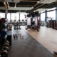 Spacious modern commercial gym space with kettlebells, dumbbell racks, cable machines, and treadmills along large floor-to-ceiling windows.