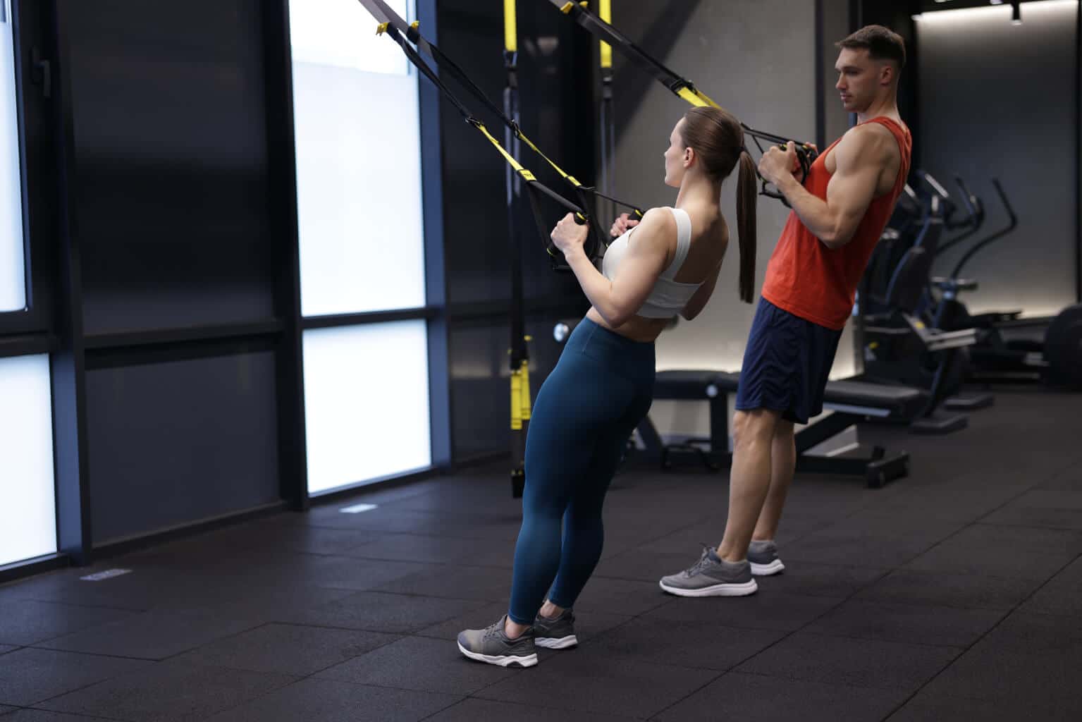A man and a woman performing bodyweight suspension training using TRX straps inside a modern gym.